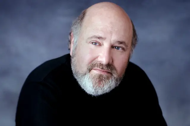 Rob Reiner is wearing a plain black long-sleeve top and is positioned against a smooth, dark gray backdrop. He has a bald head with some hair visible at the sides and a neatly trimmed gray beard. The lighting is soft and even, creating a professional studio portrait setting.
