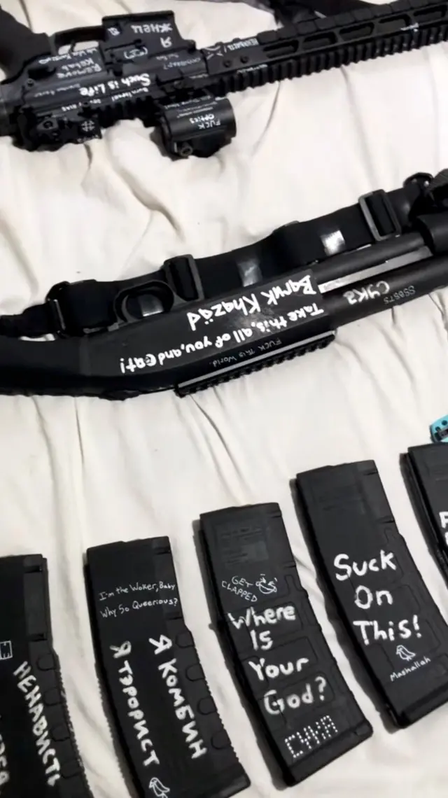 Оружие и магазины с патронами разложены на белом покрывале. На черных магазинах разные надписи белыми буквами. На одном из них по-русски написано: «Я комбин. Я террорист»