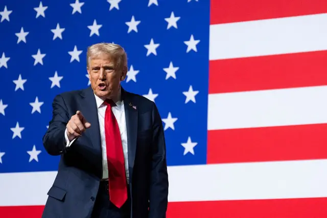 Трамп стоит на фоне американского флага и указывает пальцем в сторону зала