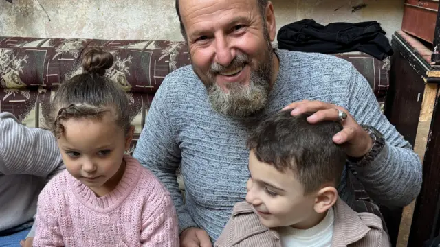 عبد اللطيف جالساً في منزله، مبتسماً، يداعب حفيديه.