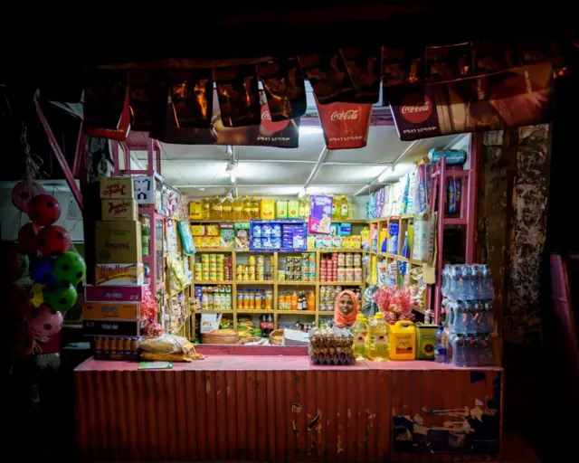 በምሽት አንዲት ባለሱቅ በትክክል ከተደረደሩ የሱቁ ሸቀጦች ጋር የሚያሳይ ምስል።