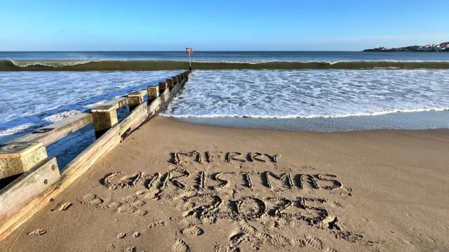 "Feliz Navidad 2025" está escrito en la arena junto a un espigón de madera. Hay pequeñas olas en la orilla y es un día soleado con cielo azul.