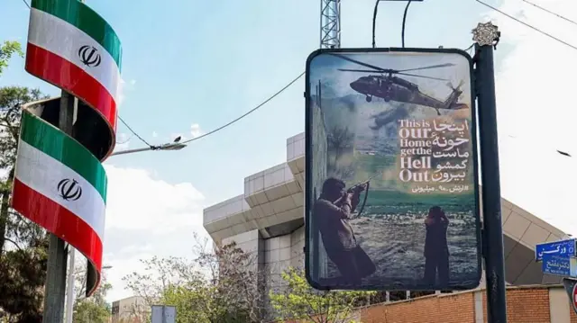 İran bayrakları sarılı bir direk ve bir başka direkteki afişte gökyüzündeki helikoptere elindeki silahla ateş açan bir erkek görülüyor ve "Burası bizim evimiz, defolup gidin" yazıyor.