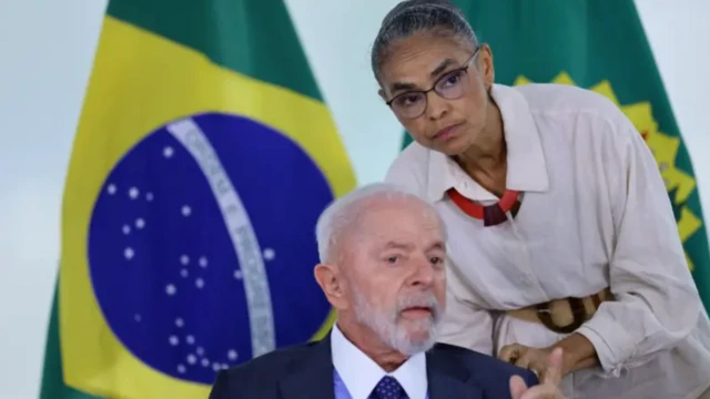 Marina Silva, de pé, fala com Lula, sentado, ambos com a bandeira do Brasil ao fundo