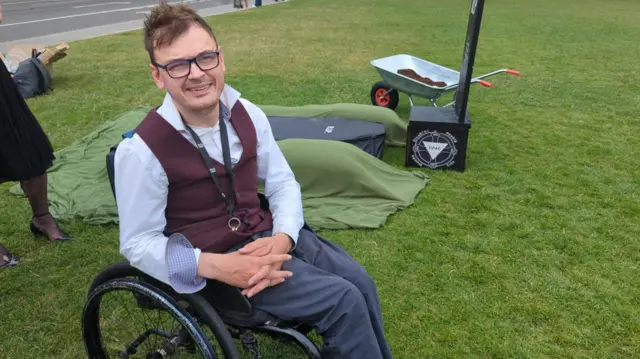 広場に作った墓の模型の横で取材に応じたフィールディング氏。車いすに座っている