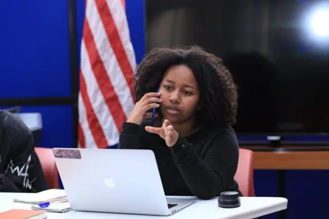 Betelhem Dessie, téléphone à l'oreille, est assise devant son PC, le drapeau américain en arrière plan. 