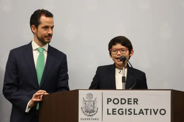 David, acompañado de un adulto, parado ante un podio y hablando al micrófono, durante una confrerencia auspiciada por el gobierno local