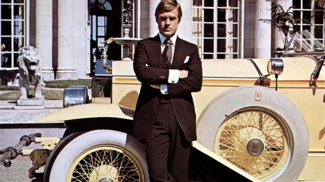 Robert Redford apparaît sur une photo promotionnelle du film Gatsby le Magnifique. Il se tient debout devant une Rolls Royce jaune vintage, les bras croisés.