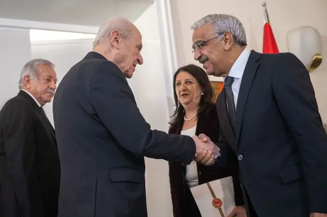 İki beyaz saçlı erkek gülümseyerek el sıkıyor. Arkada bir kadın ve bir erkek onlara bakıyor.