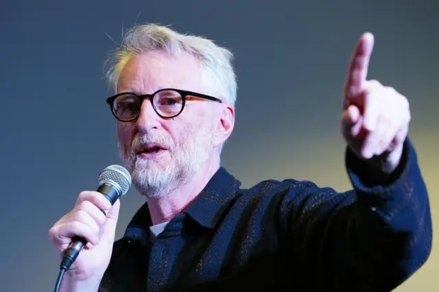 La cabeza y los hombros de Billy Bragg, con una camisa gruesa azul marino. Sostiene un micrófono en la mano derecha y apunta hacia arriba con la izquierda. Está dentro. Tiene el pelo blanco o muy rubio y barba.