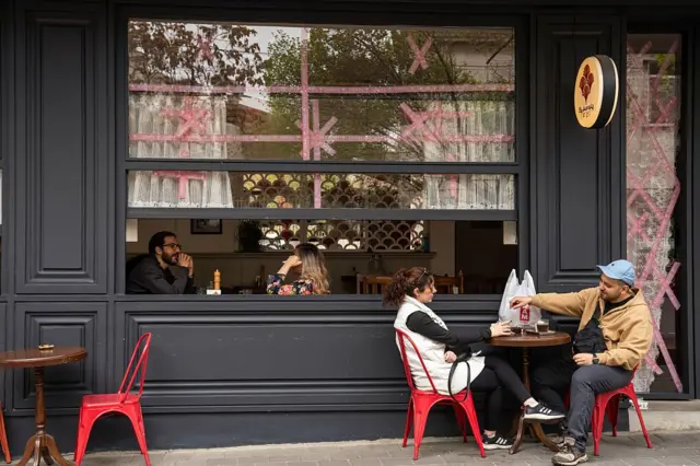 مردم در یک کافه در تهران نشسته‌اند، در حالی که برای جلوگیری از خرد شدن شیشه‌ها، روی پنجره‌ها چسب زده شده است؛ این صحنه در ۲۳ مارس ۲۰۲۶ و همزمان با ادامه حملات آمریکا و اسرائیل به ایران ثبت شده است.