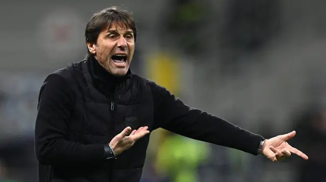 Antonio Conte in an animated pose as he points his left hand and raised his right, with his mouth open as if shouting a message. He is wearing a black top and a black gilet jacket