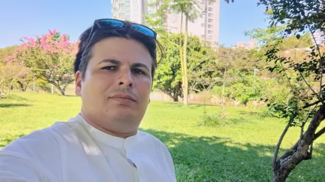 Rodrigo, vestido com uma camisa branca, olha para a câmera em selfie tirada em um jardim. No fundo, aparecem um gramado verde, árvores, e um prédio 