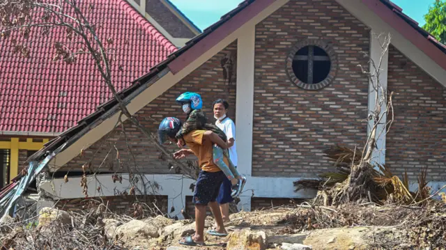 Seorang warga mengendong anaknya saat berjalan di area bencana tanah longsor di Perumahan Matauli, Pandan, Tapanuli Tengah, Sumatera Utara, Sabtu (6/12/2025). 