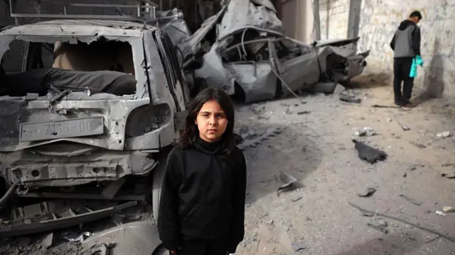 Filistinli bir kız çocuğu, 22 Nisan 2025'te İsrail ordusu tarafından yapılan hava saldırısı sırasında tahrip edilen araçların önünde siyah kapşonlu süveteriyle fotoğrafçıya bakıyor.