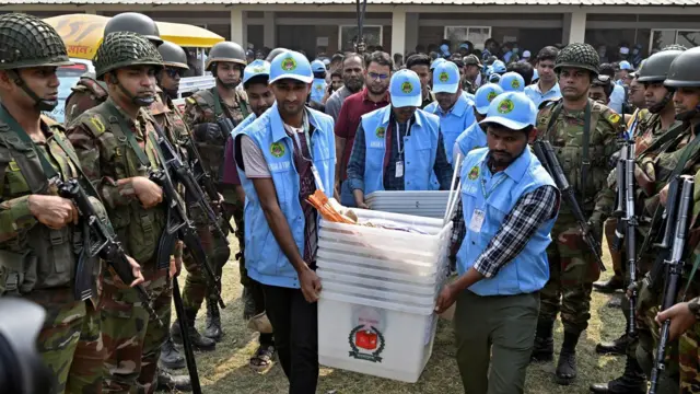 Сотрудники избиркома в Бангладеш в голубой форме несут урны для голосования сквозь строй солдат в полевой форме и с оружием
