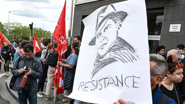 Человек на демонстрации протеста держит бумажный плакат с портретом Жана Мулена и подписью «Сопротивление». На заднем плане другие участники демонстрации с красно-белыми флагами в руках