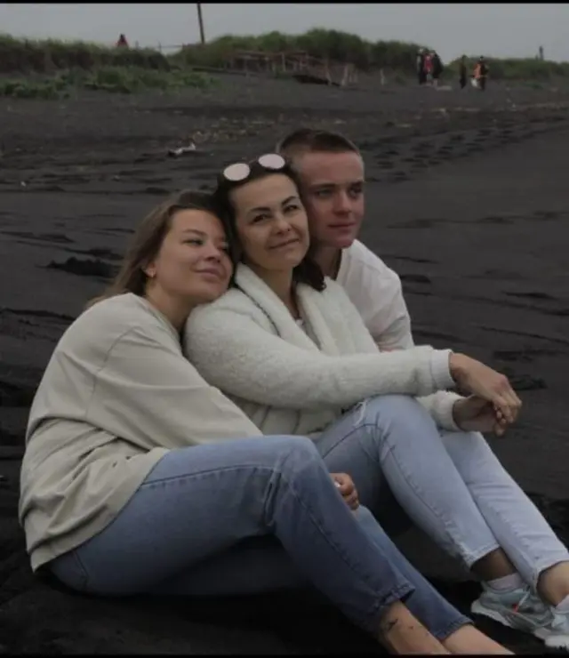 Три человека, мужчина и две женщины, в светлой одежде сидят на песке, обнявшись, и смотрят на горизонт