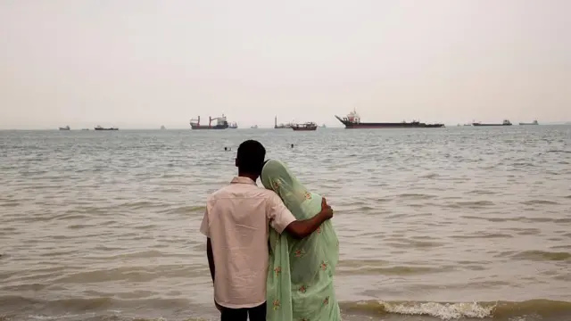 Мужчина и женщина смотрят на море, а у берега стоят на якоре корабли, 22 апреля 2026 года, Бандар-Аббас, Иран