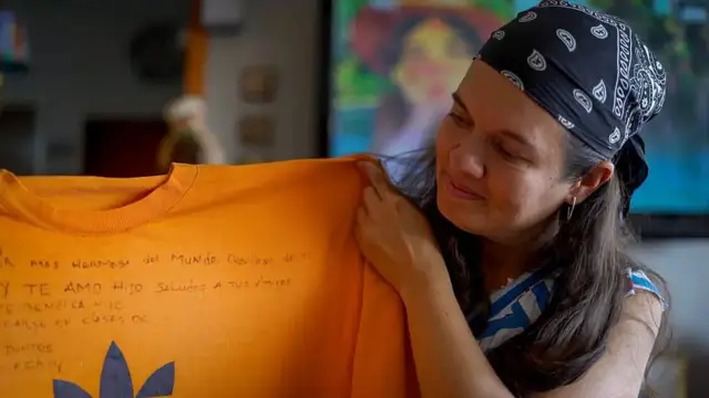 Adriana Briceño sostiene una camiseta naranja en la que hay un mensaje escrito con bolígrafo para su esposa: "Eres la mujer más hermosa del mundo. Orgulloso de ti". También hay un mensaje para su hijo: "Te amo hijo. Saludos a tus amigos. Dios te bendiga hijo. A destacarse en clases. Siempre juntos".