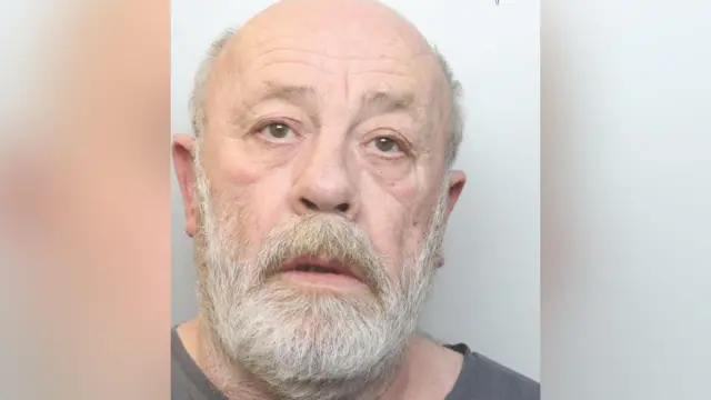 A mugshot of a bald man with a white beard and wearing a dark grey top.