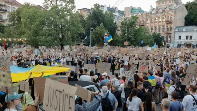Osoby trzymają tekturowe transparenty i niebiesko - żółte flagi podczas protestu w Ukrainie