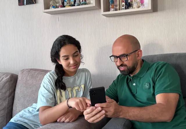 عمر يلبس قميصاً أخضر ويمسك بهاتفه ويجلس مع ابنته ليان يريها شيئاً على الهاتف