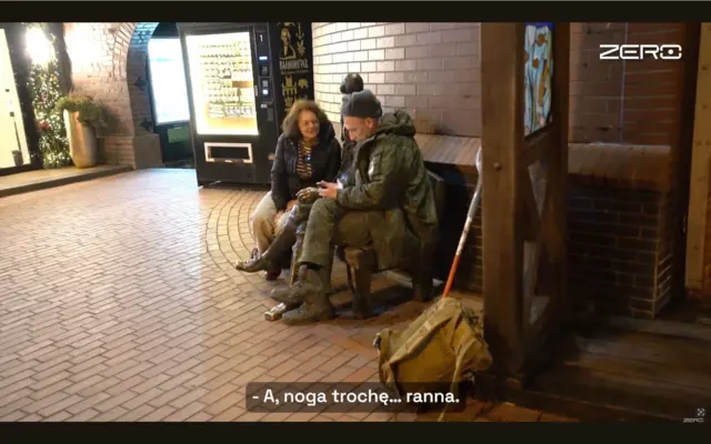 Двоє людей сидять на лавці на вулиці з бруківкою. Чоловік одягнений у камуфляжну форму й тримає в руках телефон; поруч на землі стоїть великий рюкзак та оперта тростина. Жінка сидить поруч і розмовляє з ним. Позаду видно цегляні стіни, освітлену вітрину та ялинкові гірлянди. У нижній частині зображення показано субтитр: "А, нога трохи… поранена".