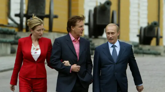 Справа наліво: Володимир Путін, Пол Маккартні в темних костюмах і його дружина Гетер Міллс у червоному діловому костюмі ідуть подвірʼям Кремля на тлі історичних гармат. 