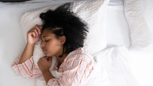 A young woman in stripey pyjamas lies in a bed asleep with her hands up to her face.