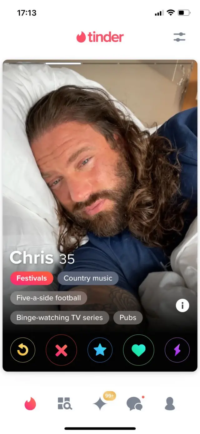 Another Tinder profile photo. It's a selfie of Harkins in bed. His head is on a white pillow. He is clothed. He has long free-flowing dark hair and a beard.