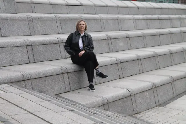 Белокурая женщина в черной кожаной куртке сидит на ступенях бетонной лестницы