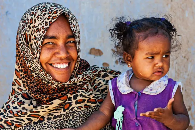 Mama wa Afrika Kaskazini aliyevaa hijabu yenye alama za duma akitabasamu akiitazama kamera, huku akiwa amemshikilia mtoto wake mchanga, ambaye amevalia nguo ya zambarau. 