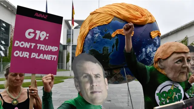 Активисты в масках Ангелы Меркель, Си Цзиньпина и Эммануэля Макрона и шар в виде Земли с прической Трампа в Берлине, июнь 2017 года. В руках у одного из активистов лозунг: G19: Don't Trump our Planet.