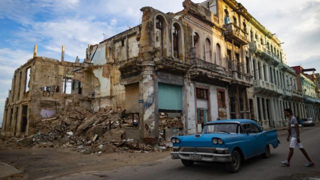 Rapaz passando por carro antigo azul em Havana, com prédio em ruínas ao fundo