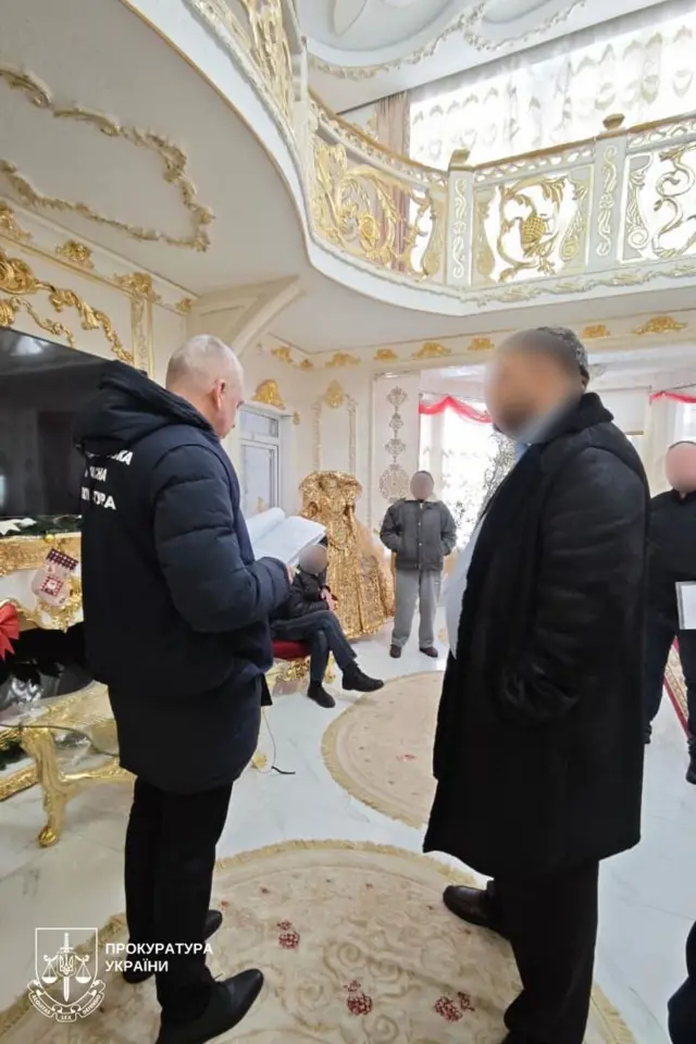 У просторому приміщенні з пишним біло‑золотим декором стоять кілька чоловіків, зайняті офіційною розмовою. Один зі співробітників у куртці читає документи. Інші учасники стоять і слухають, а один чоловік сидить у кріслі поруч. Інтер’єр вирізняється позолоченими елементами, важкими шторами та великими килимами, що створює атмосферу розкоші.