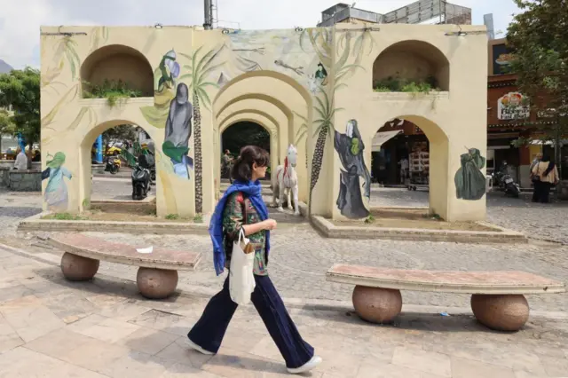 امرأة تنظر بعيداً عن الكاميرا، وتسير في أحد شوارع طهران مرتديةً الجينز وقميصاً مُلوناً بدون غطاء على رأسها، وتحمل حقيبة بيضاء .