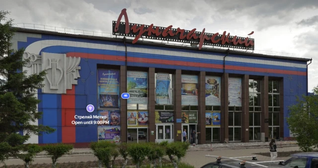 На заднем плане - серое небо. На переднем - прямоугольное здание в бело-сине-красных цветах. Слева на здании барельеф в виде духовых труб, театрального занавеса и лиры. На крыше здание вывеска в виде киноленты и красной надписи курсивом "Лунарчарский". На фасаде здания - многочисленные афиши. Перед зданием - деревья, кусты и припаркованные машины