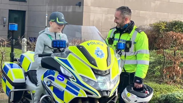 Un niño con un chándal gris está sentado en una motocicleta policial mientras un oficial de policía está de pie junto a él y le sonríe.