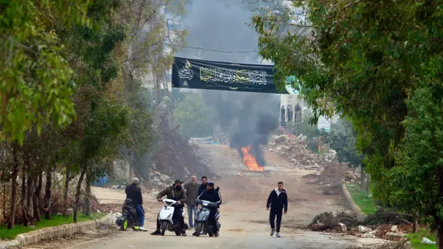 أشخاص يسيرون على طريق مدمر في جنوب لبنان، بعضهم يركب دراجات وخلفهم نيران مشتعلة، على جانبي الطريق بعض الأشجار الخضراء.