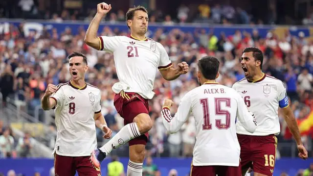 Mikel Oyarzabal jumps to celebrate scoring a penalty against France, wearing Spain's change kit of white shirt and burgundy shorts