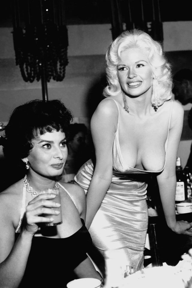 Sofía Loren, con un vestido negro con un amplio escote, está sentada y tiene un vaso en la mano. A su lado, parada y sonriendo, está Jayne Mansfield, que lleva un vestido de seda blanco con un amplio escote