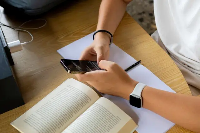 Bir çalışma masasında telefonunda mesajlaşan genç birinin elleri ve kolları. Masada kitap ve defter açık.