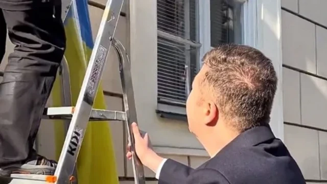 Чоловік у піджаку і коротко стрижений тримає драбину, на якій стоїть невідома людина, що знімає український синьо-жовтий прапор із будівлі