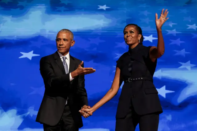 Barack e Michelle Obamasite de aposta cartolaum fundo azul: ele estásite de aposta cartolaterno preto com uma gravata azul, segurando a mão direita da esposa, que está vestida com um elegante conjunto preto.