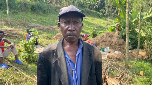 Umugabo urimo kureba muri 'camera'. Yambaye ikoti ry'umukara n'ishati y'ubururu. Iruhande rwe inyuma hari abagabo bicaye mu byatsi, inyuma ye hari abagore bunamye begeranye n'ibishishwa by'ibishyimbo byahuwe. Iruhande rw'abo bagore hari insina. 