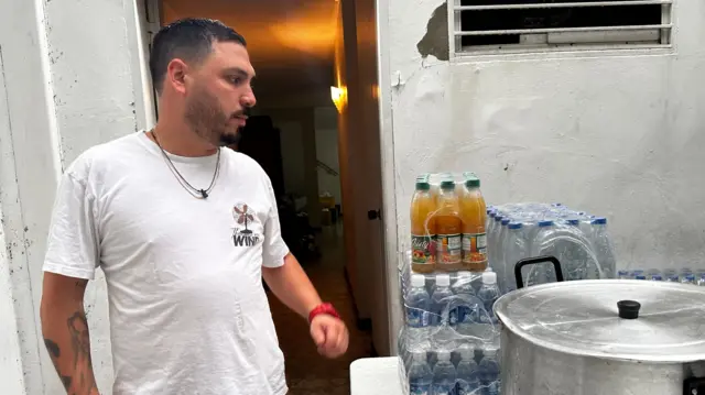 Víctor Andrea D'Orsi en el patio al lado de una olla y botellas de agua. Lleva una franela blanca.