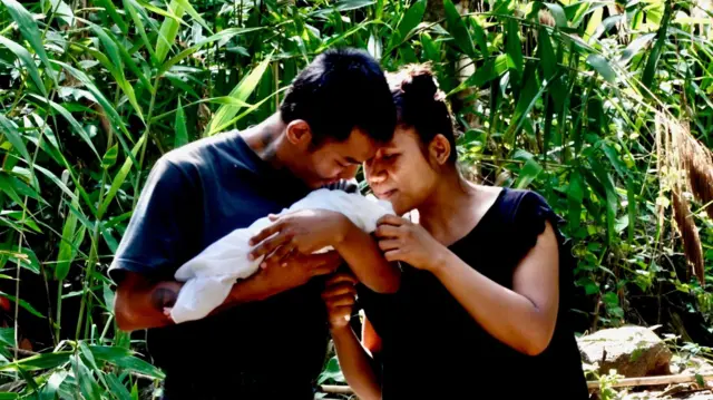 A couple hold a newborn baby in the arms, standing in front of the green of the jungle
