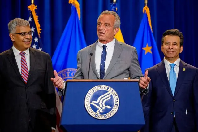 Three men, all wearing suits and ties, stand on a stage. The man in the middle stands behind a podium bearing the stamp of the Department of Health and Human Services. He is talking and gesticulating with his hands. The other two men are smiling.