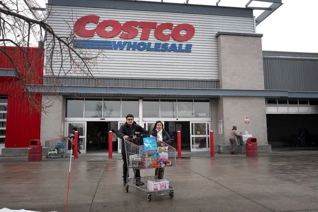 Una pareja empuja un carrito de supermercado a las afueras de un edificio con un gran letrero con letras rojas que dice Costco.